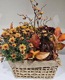 Autumn fall floral arrangement in a woven basket with yellow and orange chrysanthemums, glass pumpkin lantern, decorative leaves, and berry branches.